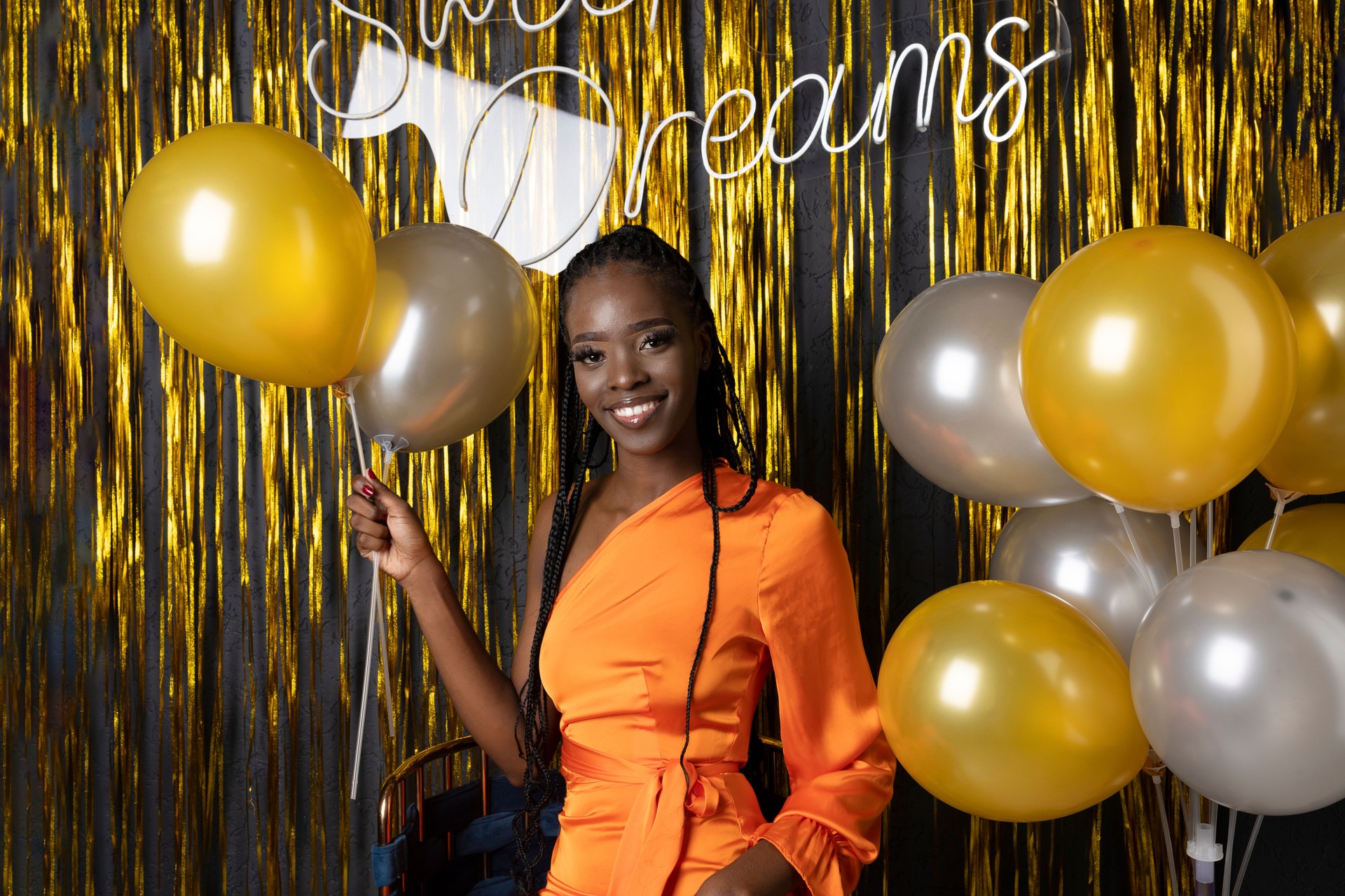 Portrait of charming black woman in elegant formal dress in festive hall with gold decorations and colored balloons. Joyful birthday lady in photo zone. Birthday party, showy holiday, celebration